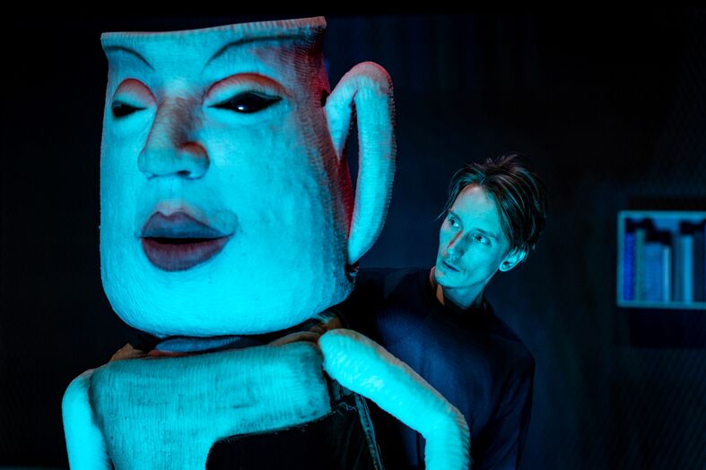 Een acteur interacteert met een grote, kleurige pop in een theatrale setting met gedimde lichten.