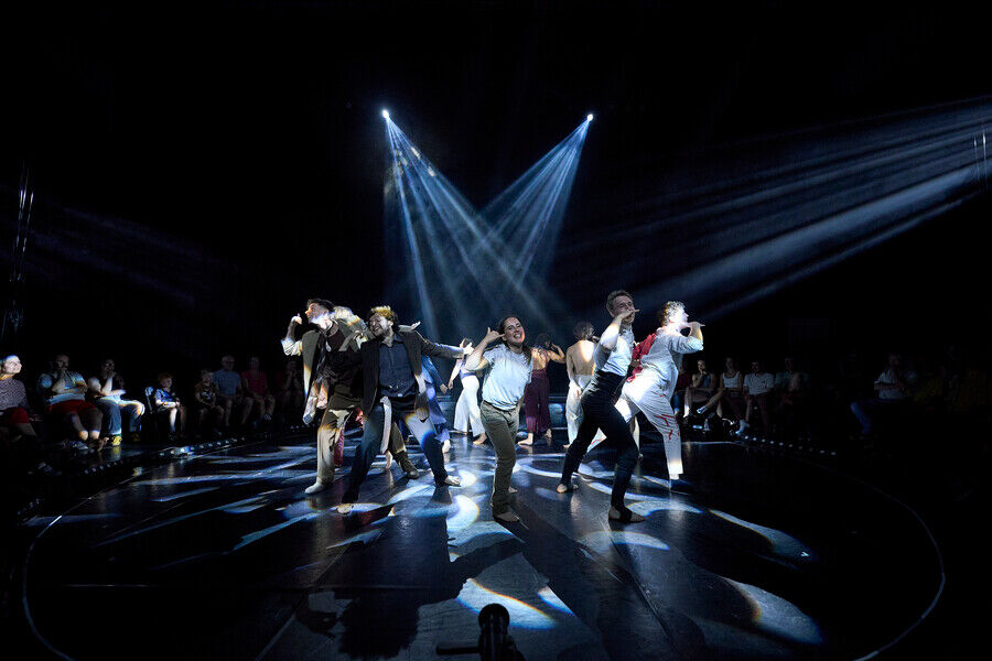 Groep performers voert een energieke choreografie uit op een podium, verlicht door fel licht. Publiek kijkt toe.