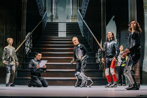 Vijf acteurs in historische kostuums staan op een trap, terwijl één acteur een script leest.
