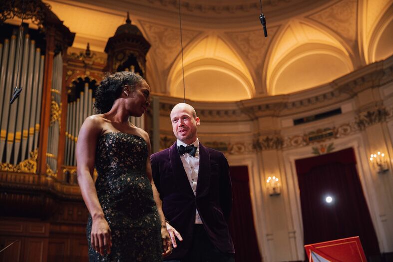 Een man en een vrouw in formele kleding staan op een podium in een elegante zaal met een orgel op de achtergrond.