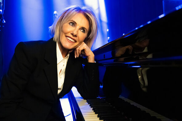 Vrouw in een zwart pak glimlacht terwijl ze leunt op een piano, gefotografeerd op een podium met blauwe verlichting.