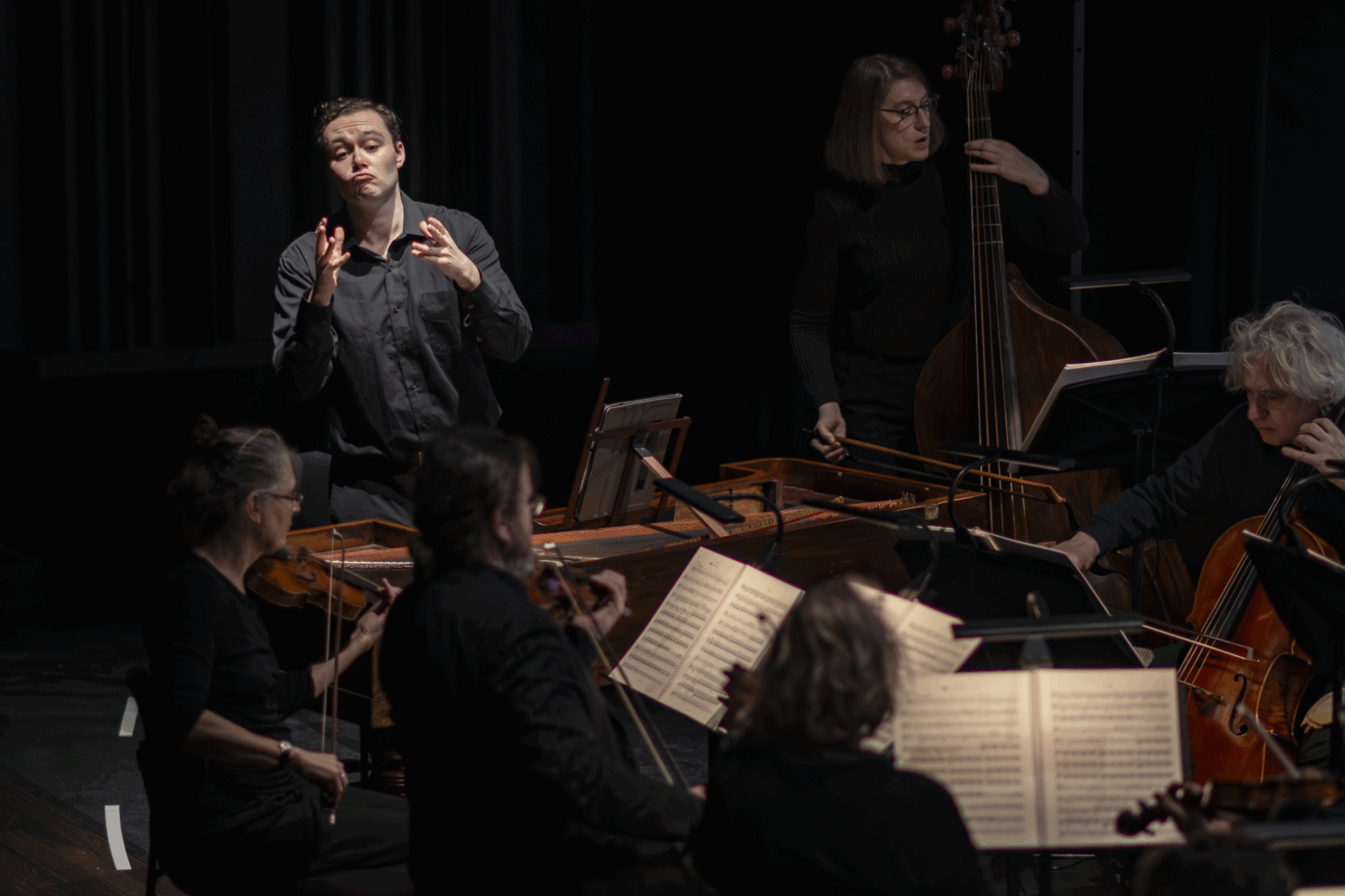 Dirigent dirigeert een ensemble van muzikanten in een donkere concertzaal. Instrumenten zijn zichtbaar, evenals bladmuziek.