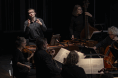 Dirigent dirigeert een ensemble van muzikanten in een donkere concertzaal. Instrumenten zijn zichtbaar, evenals bladmuziek.