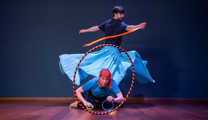Twee performers bewegen met hoepels, één draaiend met rok en lint, één laag bij vloer.