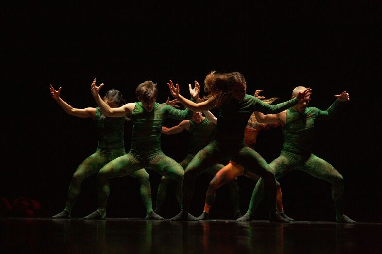 Een groep dansers in groene bodystockings voert een expressieve dans uit op een donkere achtergrond.