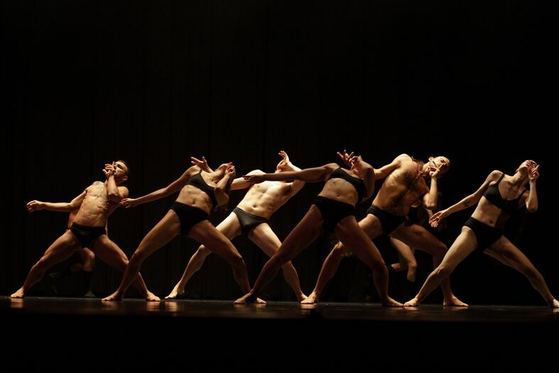 Zeven dansers in zwart ondergoed voeren een dynamische dansroutine uit op een donkere achtergrond.