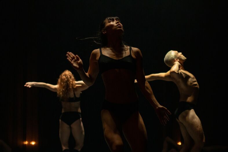 Drie dansers in ondergoed voeren een expressieve dansvoorstelling uit op een donker podium met spotlights.