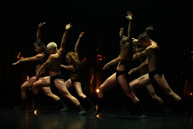 Zes dansers in zwart ondergoed voeren een energieke choreografie uit op een donker podium met sfeervolle belichting.