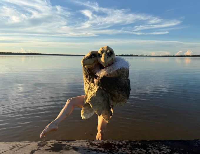Dansende persoon in een roofvogel kostuum bij de oever van een rustig water, met een heldere lucht op de achtergrond.
