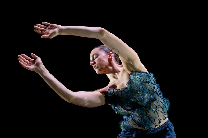 Een danseres in een elegante pose met uitgestrekte armen, gekleed in een stijlvol, doorzichtig topje tegen een zwarte achtergrond.