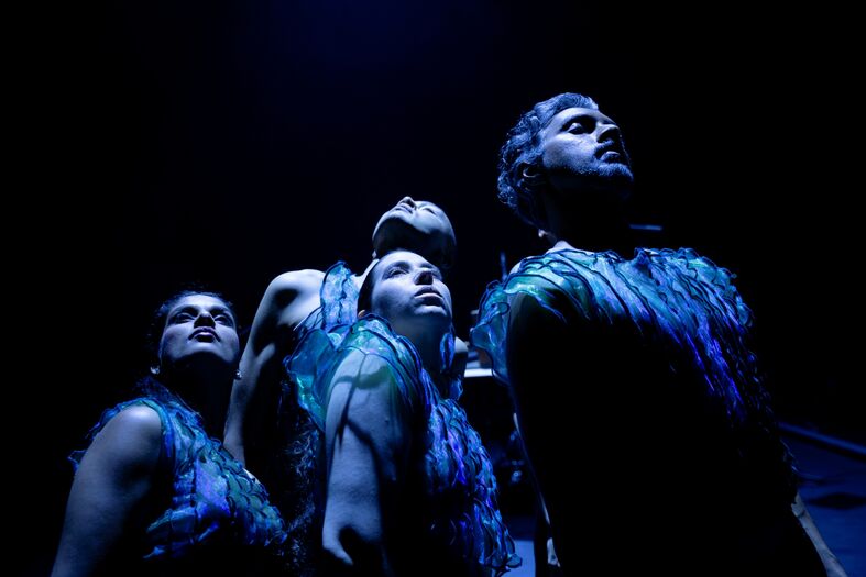 Groep artiesten in donkere setting, kijkend omhoog met theatrale outfits en blauwe verlichting.
