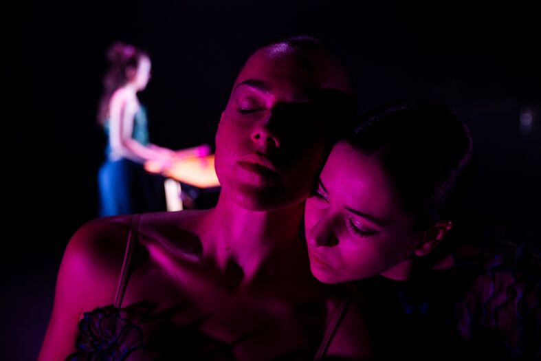 Twee dansers staan dicht bij elkaar in een sfeervol verlichte ruimte met paarse en roze tinten.
