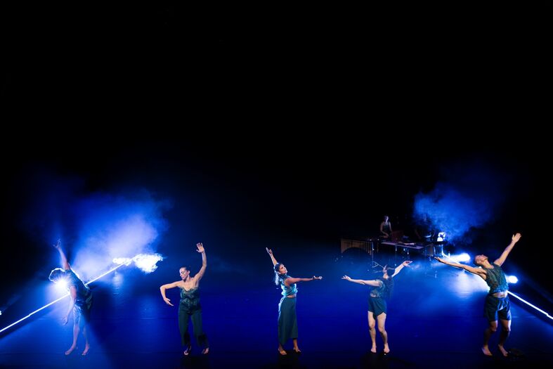 Zes dansers voeren een choreografie uit op een podium met blauwe verlichting en een donkere achtergrond.