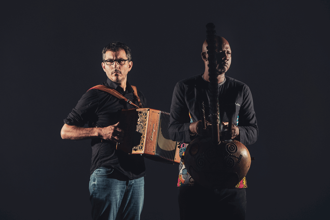 Twee muzikanten staan zij aan zij, één met een accordeon en de ander met een Afrikaanse snaarinstrument.