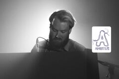 Persoon met baard zit achter piano en microfoon in zwart‑wit tegenlicht.