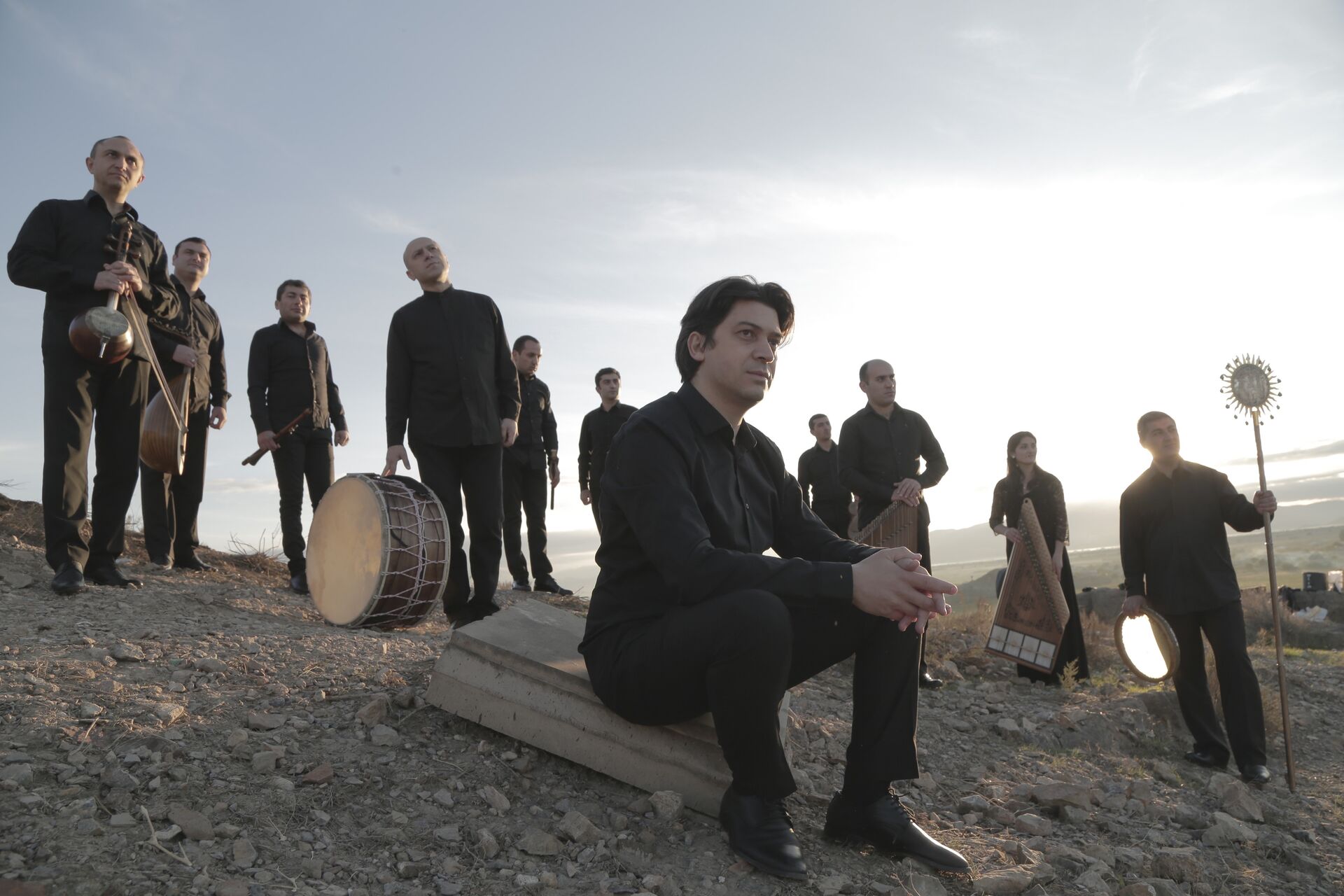 Groep muzikanten poseert op een verhoogd terrein met instrumenten bij zonsondergang. Een man zit vooraan.