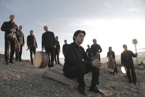 Groep muzikanten poseert op een verhoogd terrein met instrumenten bij zonsondergang. Een man zit vooraan.
