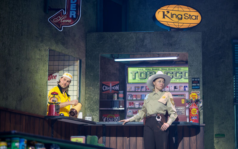 Twee acteurs in een winkelsetting, één in een gele outfit achter de toonbank, de ander in een cowboykostuum voor de toonbank.