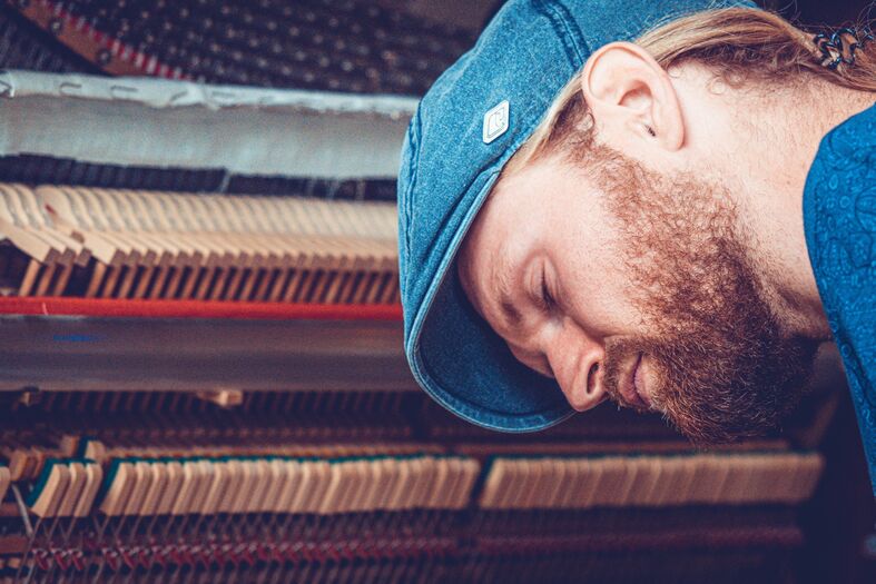 Man met een baard en pet, leunt voorover terwijl hij een piano inspecteert.