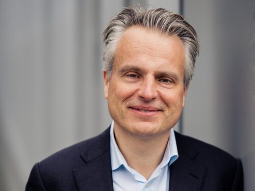 Een man met grijs haar in een zwarte blazer stelt zich glimlachend voor tegen een neutrale achtergrond.