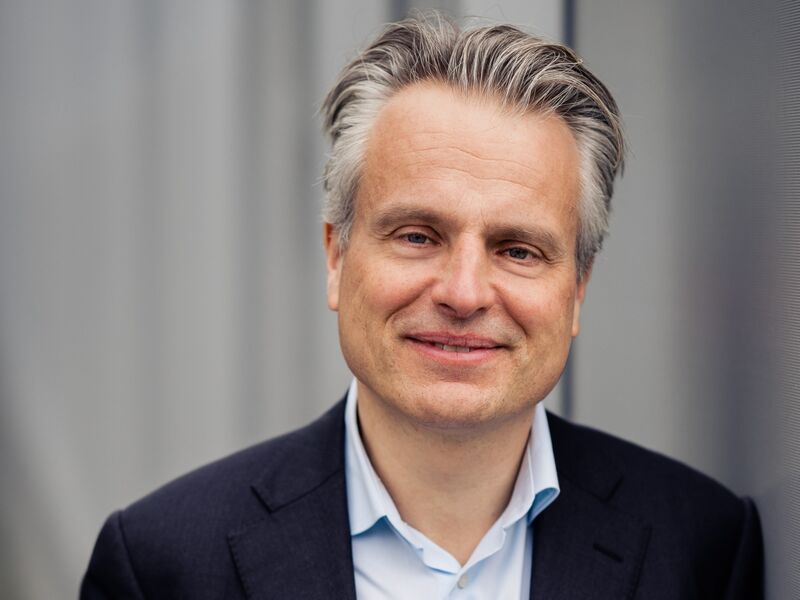 Een man met grijs haar in een zwarte blazer stelt zich glimlachend voor tegen een neutrale achtergrond.