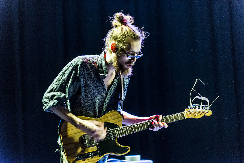 Man met een baard speelt gitaar op een podium, met een donkere achtergrond en lichteffecten.