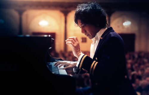 Concertpianist speelt op een vleugelpiano, gekleed in elegante outfit, met publiek op de achtergrond.