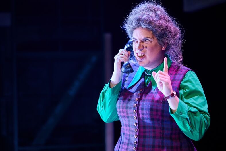 Vrouw in kostuum met een telefoon op het oor, in een theatrale setting, spreekt met nadruk en bezorgdheid.