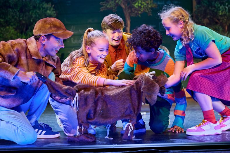 Groep kinderen op een podium, vrolijk bezig met een bruine hond tijdens een theateruitvoering.