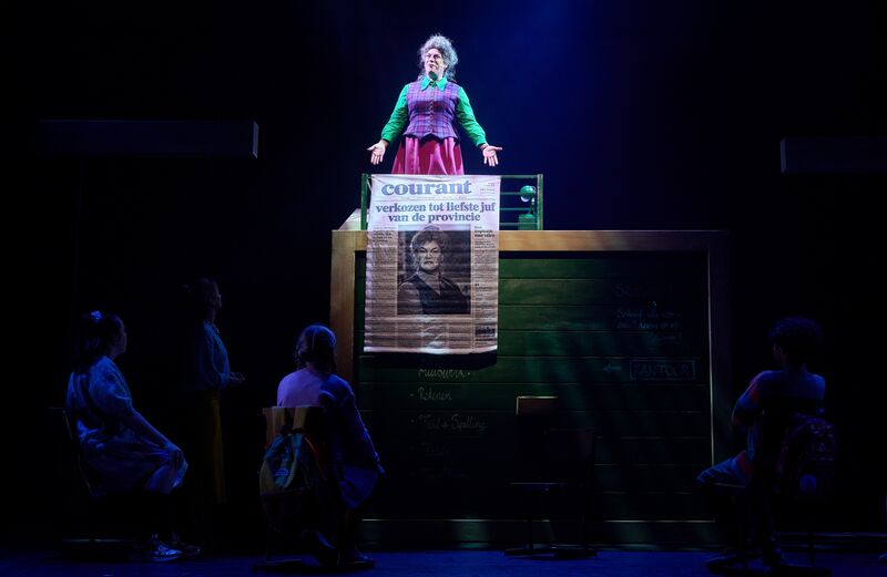 Actrice in kleurrijk kostuum staat op een podium met een spandoek, omringd door andere performers in een theaterproductie.