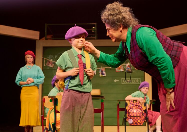 Twee actrices interactieren op een podium, terwijl een jongen in kleurrijke kledij en hoed zich voorbereidt op een optreden.