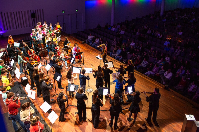 Een orkest voert een concert uit op het podium voor een publiek in een zaal met kleurrijke verlichting.
