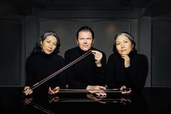 Drie muzikanten in zwarte kleding, zittend aan een tafel met strijkinstrumenten in hun handen, tegen een neutrale achtergrond.