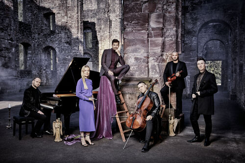 Een groep van zes muzikanten poseren creatief in een historische, ruïneachtige setting met diverse instrumenten.