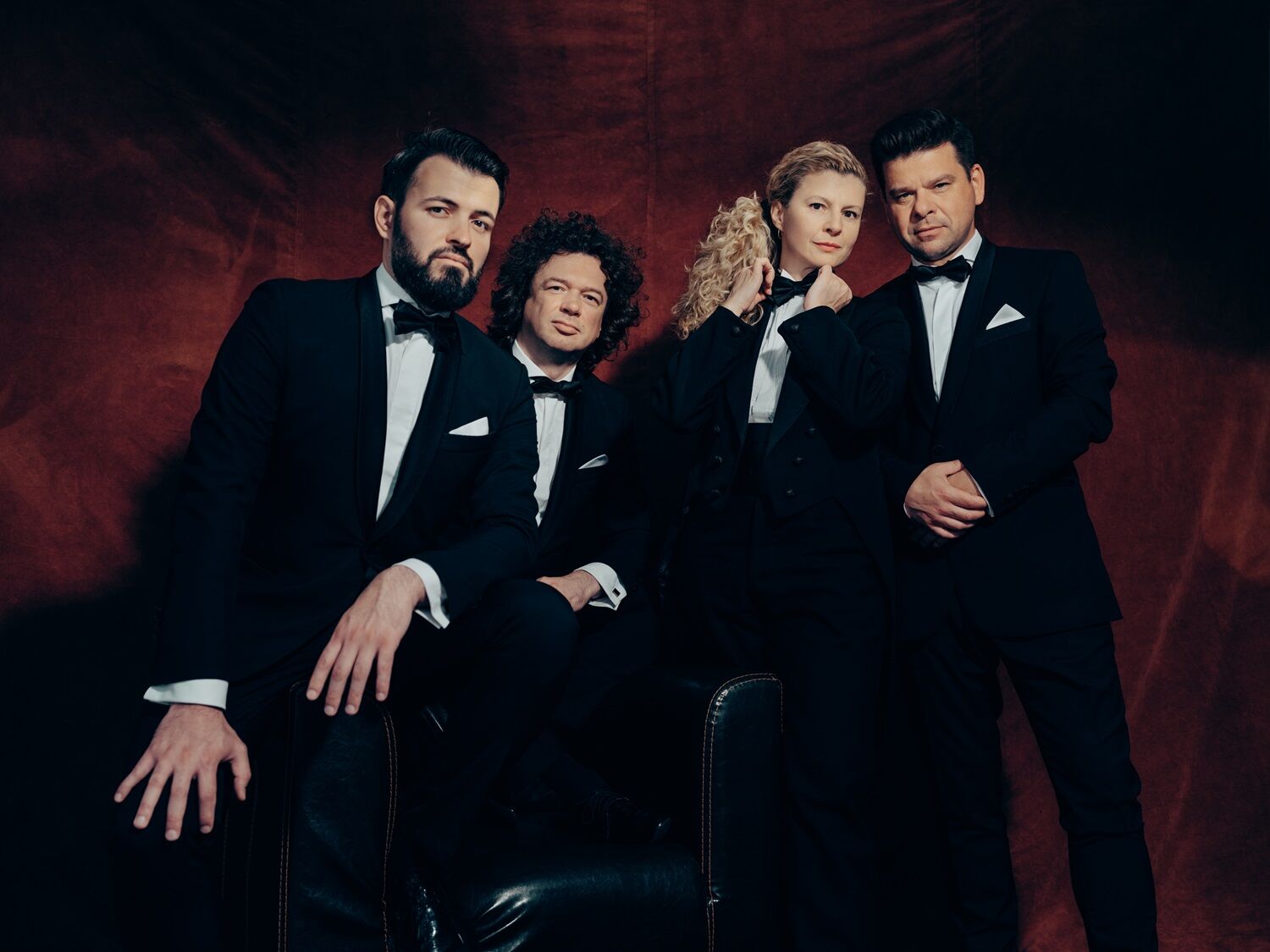 Vier mensen in tuxedo's poseren serieus voor een rode achtergrond, met een mix van krachtige en elegante houdingen.