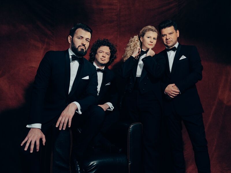 Vier mensen in tuxedo's poseren serieus voor een rode achtergrond, met een mix van krachtige en elegante houdingen.