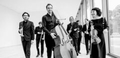 Groep muzikanten in zwart-witte kleding poseert samen in een moderne ruimte, met diverse instrumenten in de hand.