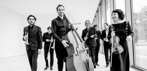 Groep muzikanten in zwart-witte kleding poseert samen in een moderne ruimte, met diverse instrumenten in de hand.