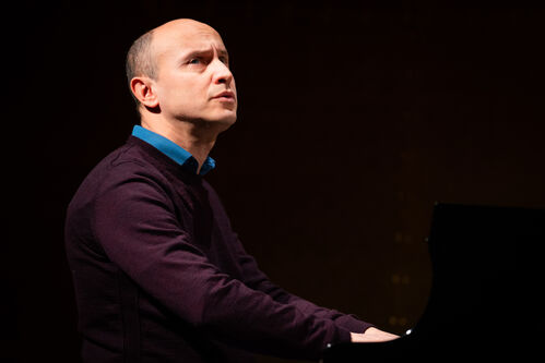 Man met een donker paarse trui speelt piano, terwijl hij omhoog kijkt naar het publiek op een podium.