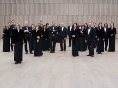 Groepsportret van een symfonieorkest, gekleed in formele zwarte kleding, staand in een moderne concerthal.