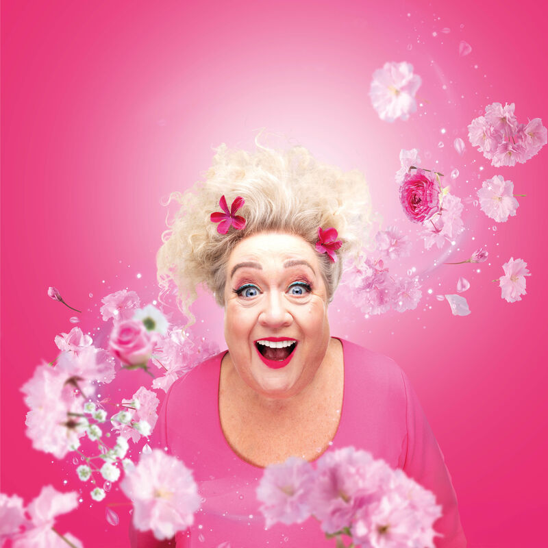 Christel de Laat met een grote, blijde lach, omringd door roze bloemen en een roze achtergrond.