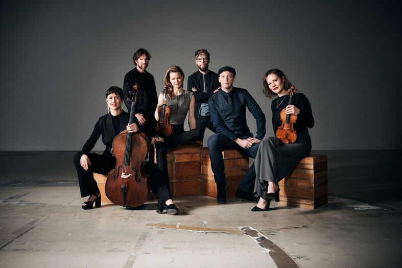 Zes muzikanten poseren samen in een studio, met strijkinstrumenten en een neutrale achtergrond.