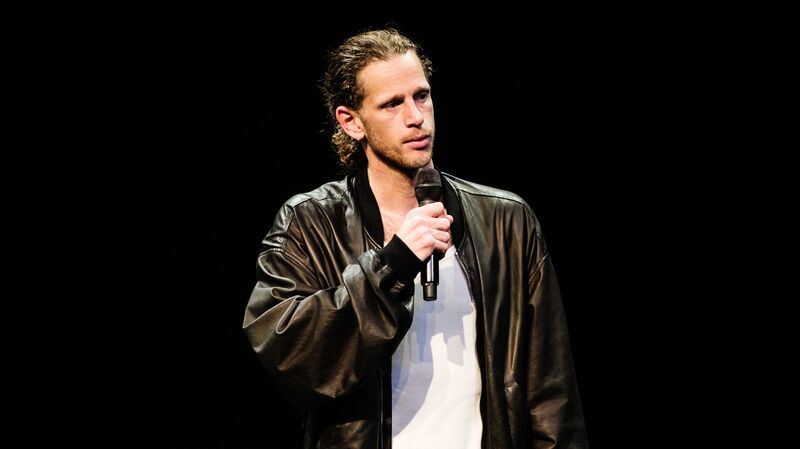 Man met een mic in een zwart leren jack, staat op een podium met een donkere achtergrond.