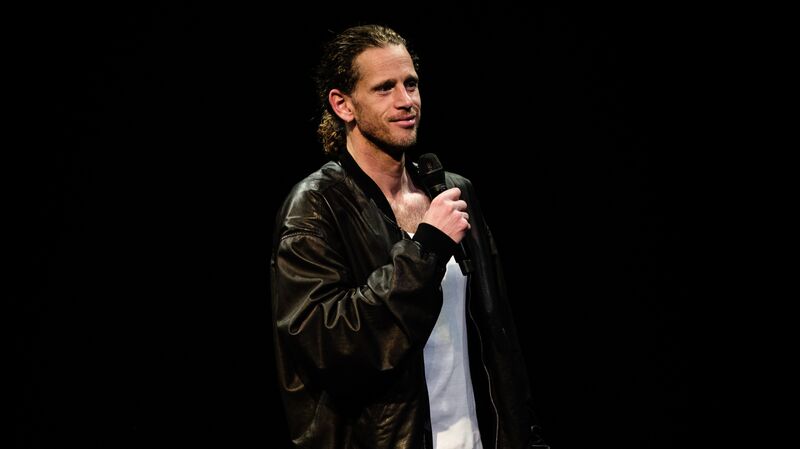 Man met een microfoon op een podium, gekleed in een zwarte leren jas en witte T-shirt, staande in een donkere omgeving.