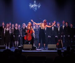 Een zangperformantie met een koor op het podium, met een cellist en een zangeres in de voorgrond, onder een kroonluchter.