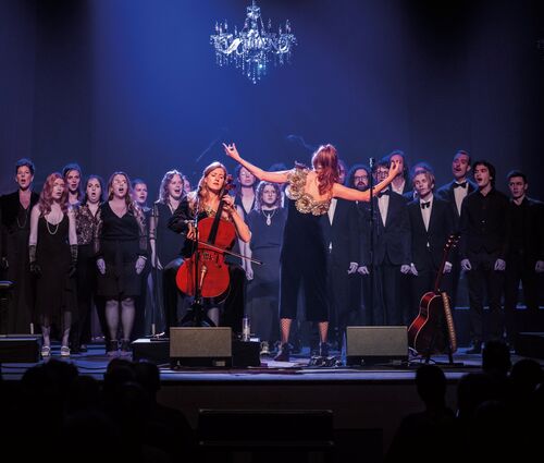 Een zangperformantie met een koor op het podium, met een cellist en een zangeres in de voorgrond, onder een kroonluchter.