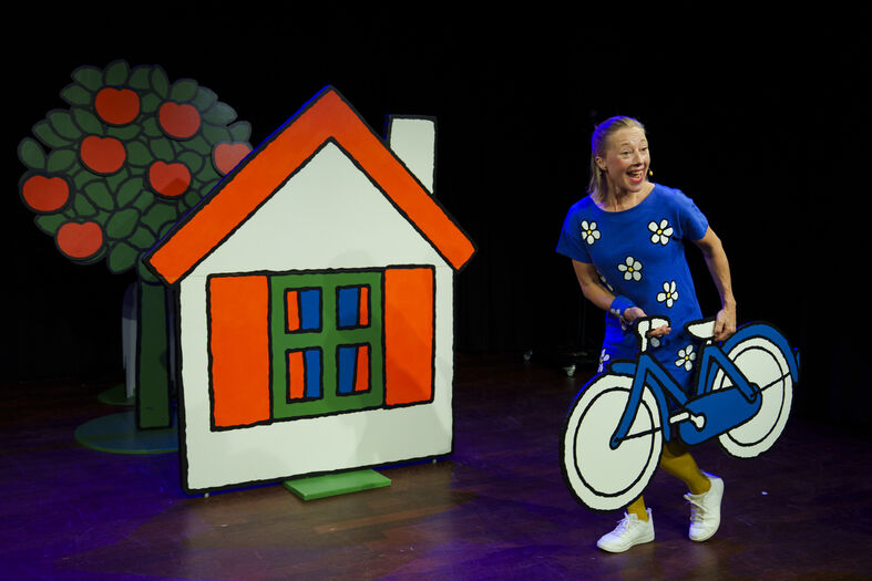 Een vrouw in een blauwe jurk houdt een grote kartonnen fiets vast, naast een kleurig huis en een boom.