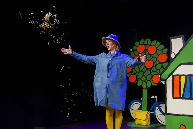 Een vrolijke performer in een blauwe outfit staat op een podium met een kleurrijk decor van een huis en een boom.