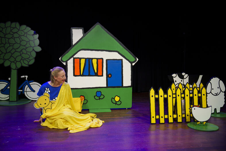 Een actrice in een gele jurk zit naast een gekleurde poppenhuis met een hek en dieren op het podium.