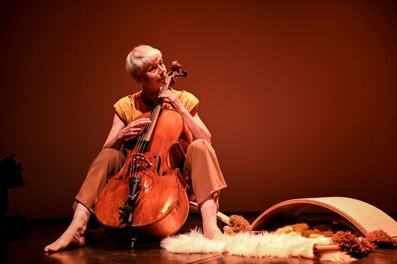Een muzikante met kort haar speelt cello op een podium, omringd door kleurrijke objecten en zachte stoffen.